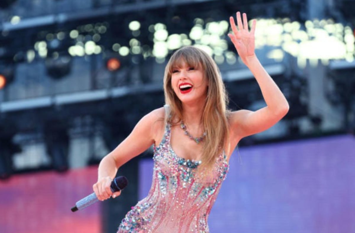 A smiling woman with long hair wearing a sparkling, colorful dress holds a microphone and waves while performing on an outdoor stage with blurred lights in the background.