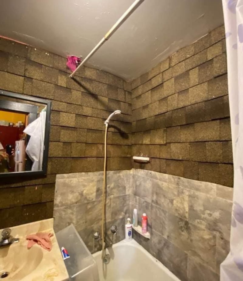 A bathroom with a bathtub, showerhead, and beige sink. The upper walls are covered in brown roof shingles, while the lower walls have gray tiles. Toiletries are placed on the tub edge and a shower curtain is partly visible.