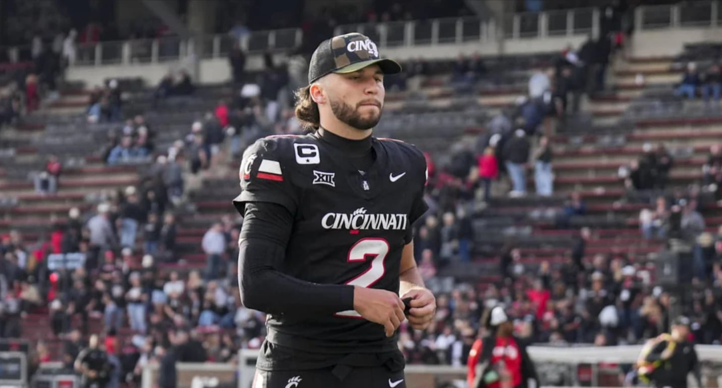 A football player in a black Cincinnati Bearcats uniform and cap stands on the field with a focused expression, as people in the stadium seating are blurred in the background.