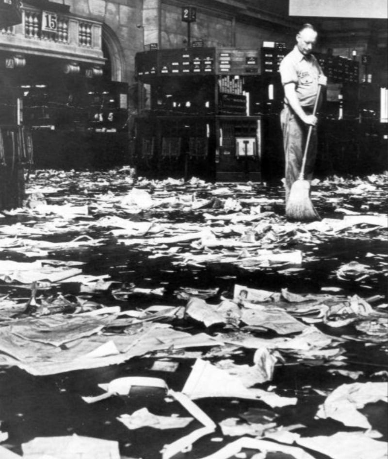 A man sweeps a large floor covered with scattered papers and debris inside a spacious room with rows of desks and high ceilings, creating a sense of disorder and aftermath.