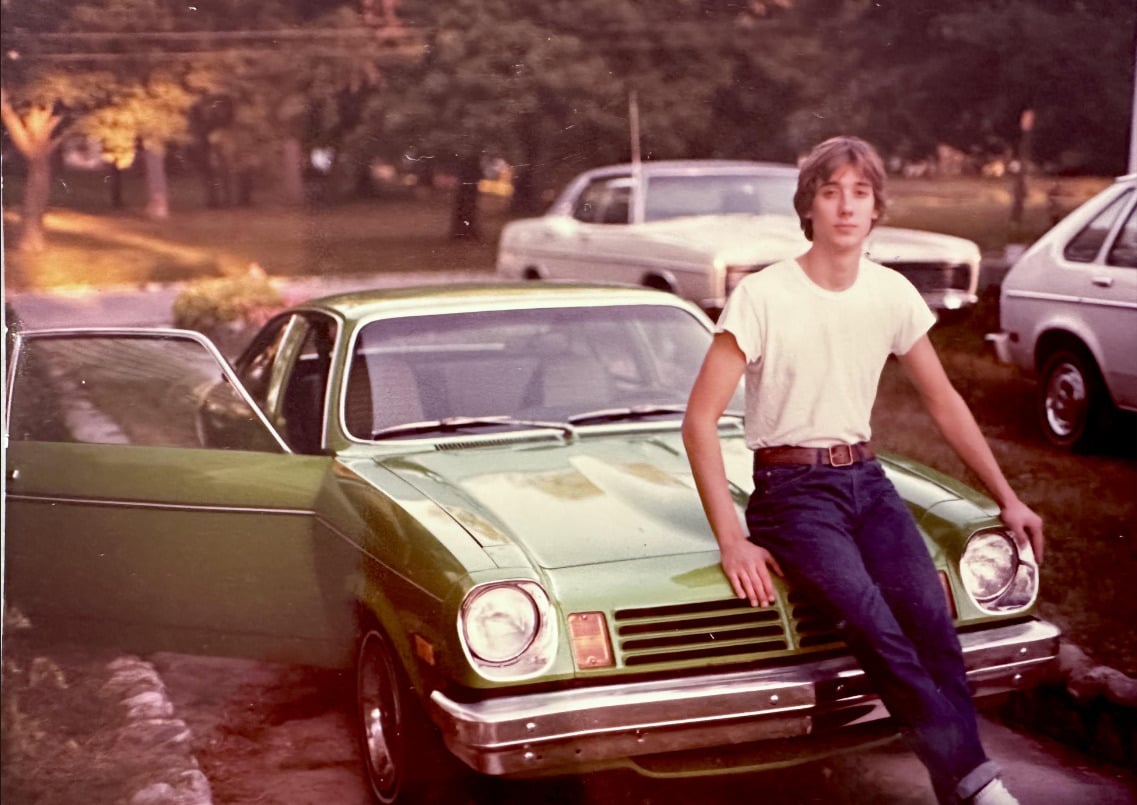 A young person in a white T-shirt and jeans sits on the hood of a green vintage car with the driver’s door open. Two older white cars are parked behind, with trees and grass in the background.