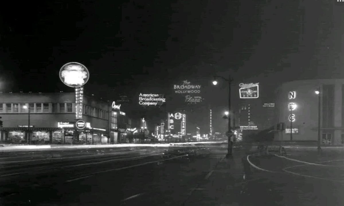 A black-and-white nighttime photo of a city street lined with illuminated neon signs, including NBC, American Broadcasting Company, and various theaters. The street appears mostly empty, with buildings and streetlights visible.
