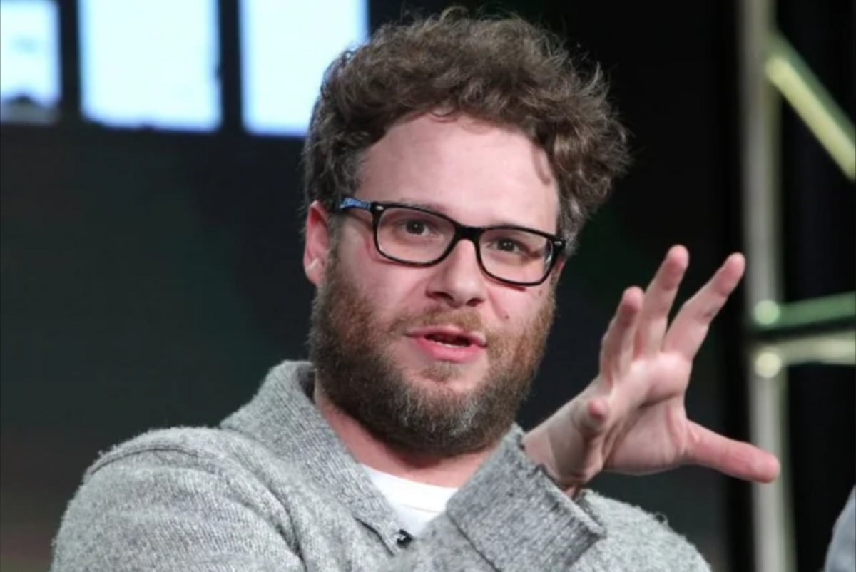 A bearded man with curly hair and glasses, wearing a gray sweater, gestures with his hand while speaking onstage against a dark background with blurred lights.