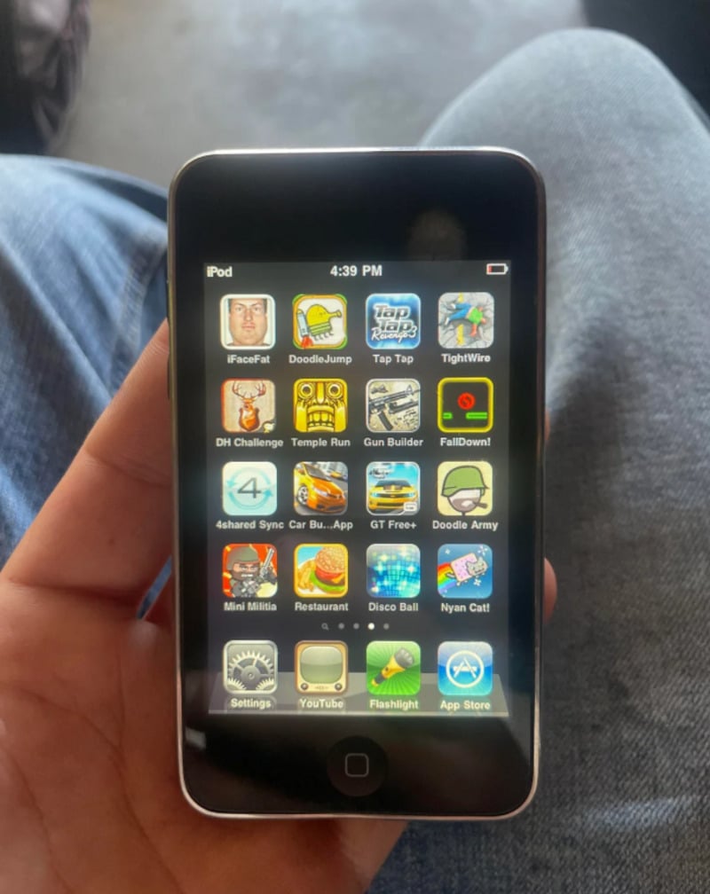 A hand holds an old iPod Touch displaying its home screen, which shows various classic app icons and games like Doodle Jump, Temple Run, Tap Tap, and YouTube. Blue jeans are visible in the background.