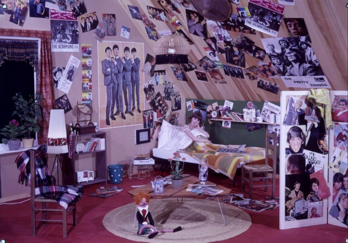 A cozy attic bedroom is filled with Beatles and other 1960s band posters, records, and memorabilia. A girl sits on her bed reading, while a doll is on the floor. The room is colorful and cluttered with books and decor.