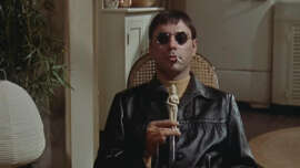 A man in dark sunglasses and a black leather jacket sits on a chair, holding a small statue and smoking a cigarette, in a room with plants and shelves in the background.