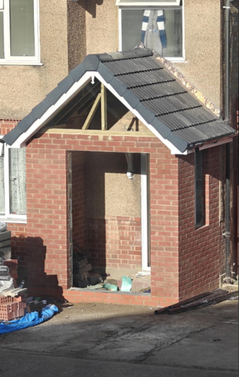 A small brick porch with a triangular roof is under construction in front of a house. The structure has no door or windows installed yet, and building materials are scattered inside and nearby.