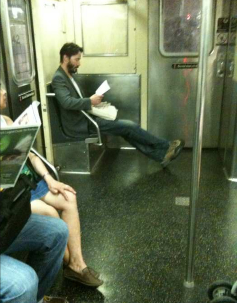 A man with a beard and casual clothes sits alone on a subway bench, reading papers with his legs stretched out across the empty seats. Another person sits nearby, reading a newspaper. The subway car appears quiet.