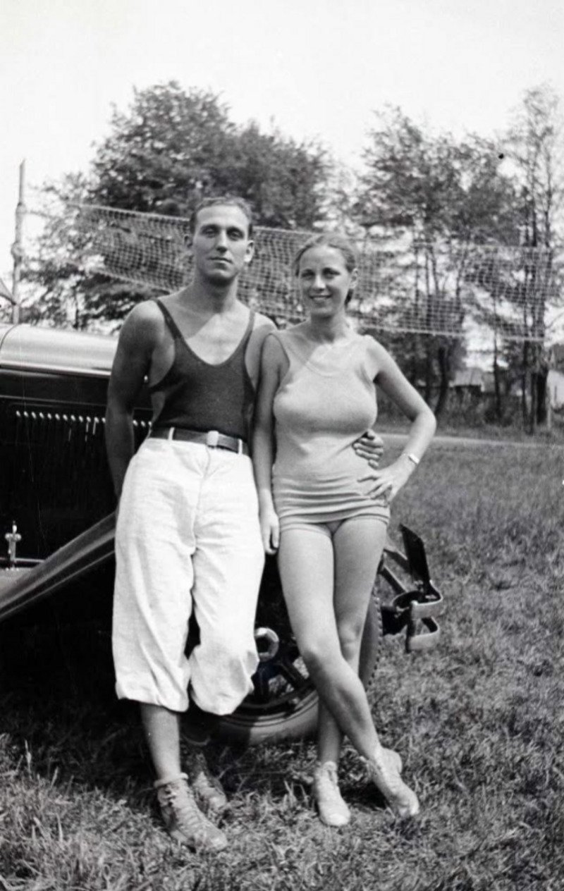 A man in a tank top and rolled-up pants stands next to a woman in a vintage swimsuit, both smiling beside an old car on a grassy field with trees and a net in the background.