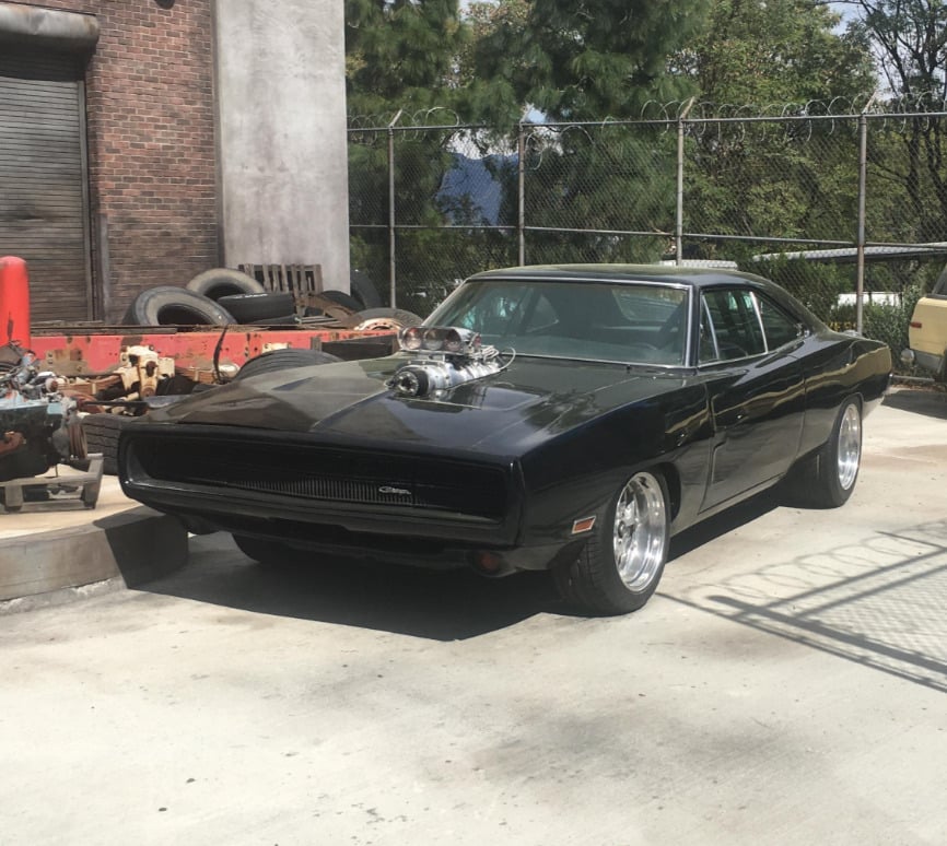 A classic black muscle car with a supercharger protruding from its hood is parked on a concrete surface near a chain-link fence, with trees and industrial equipment in the background.