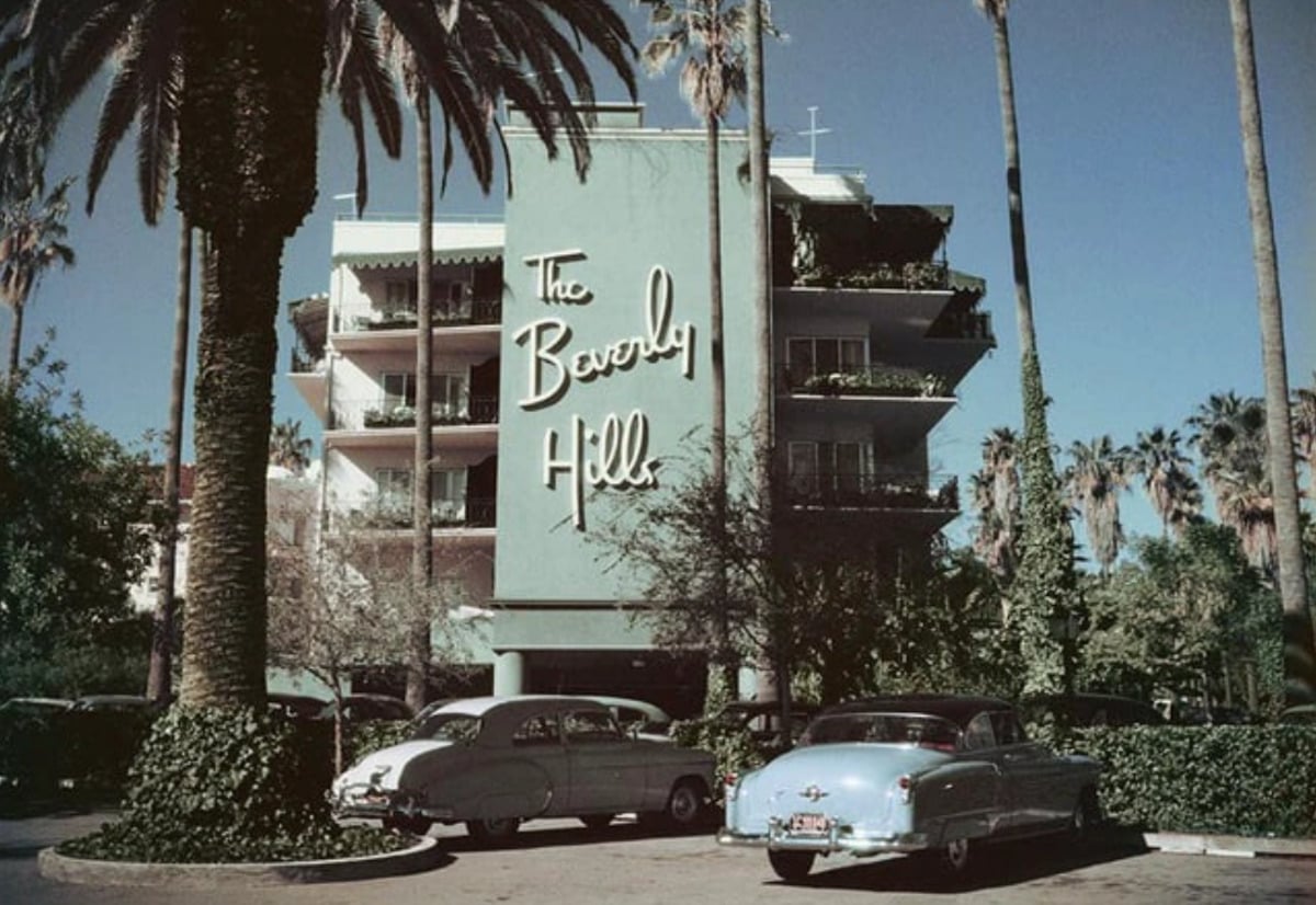A vintage photo of The Beverly Hills Hotel shows classic cars parked in front, tall palm trees, and the building’s iconic sign in cursive lettering on a green facade. The scene has a retro, mid-20th-century feel.