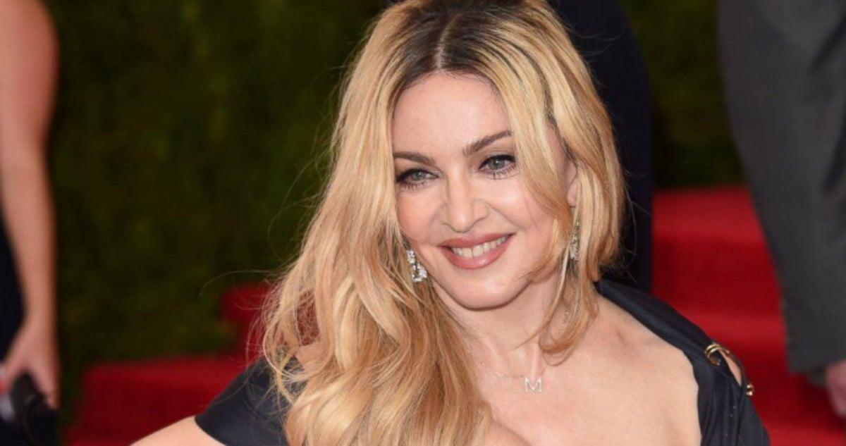A woman with long, wavy blonde hair smiles at the camera, wearing a black dress with a plunging neckline, drop earrings, and a necklace with an "M" pendant. The background features red carpeted stairs and greenery.
