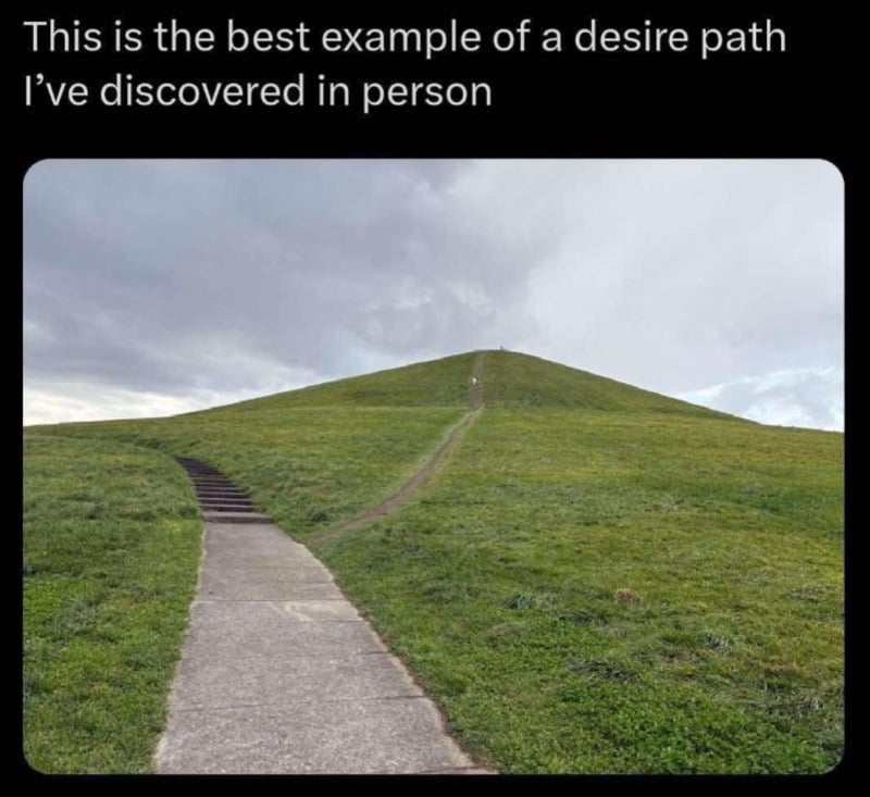 A paved sidewalk curves left around a grassy hill, but a worn dirt path cuts straight up the hill, showing where people have walked directly instead of following the sidewalk.