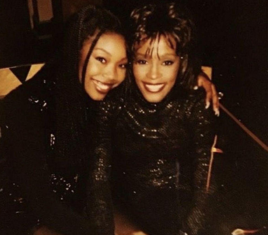 Two women sit closely together, smiling at the camera. Both wear sparkly black outfits, and one has braided hair while the other has short, styled hair. They appear happy and relaxed in a warmly lit setting.
