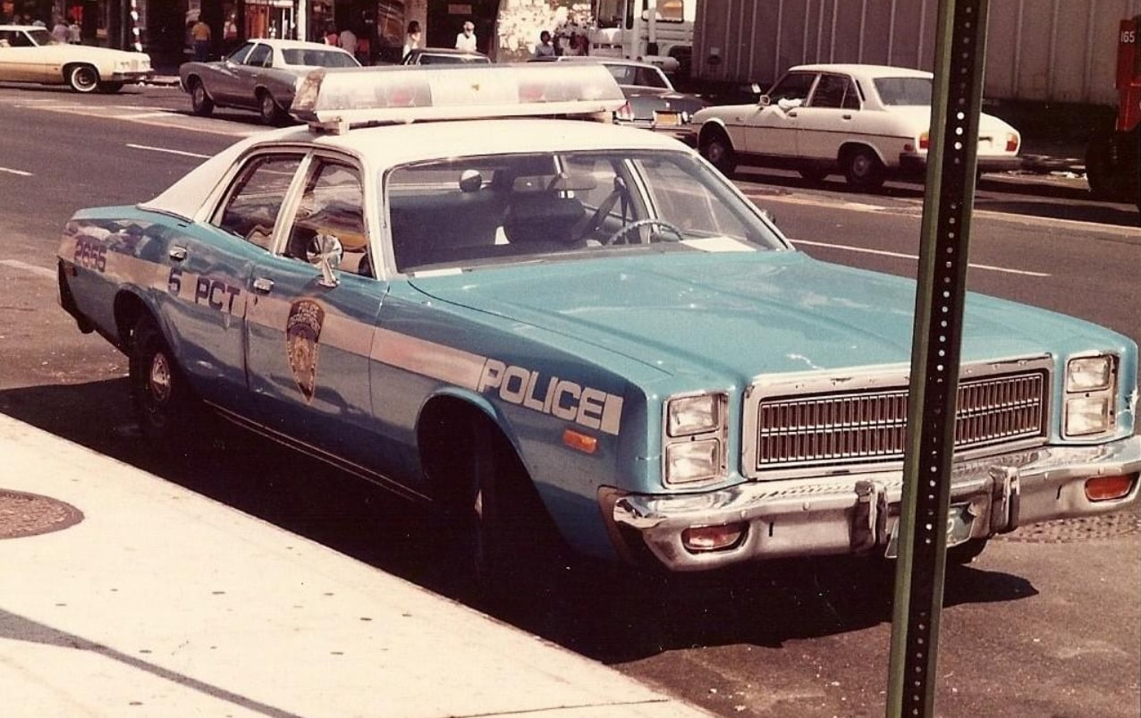 A vintage blue and white police car is parked on a city street. The car features "POLICE" and "6 PCT" markings and has a red and blue light bar on the roof. Other vehicles are visible in the background.