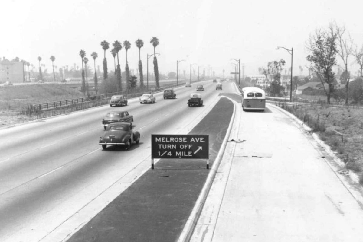 Black-and-white photo of a 1940s freeway with vintage cars and a bus. A sign on the median reads, "MELROSE AVE. TURN OFF 1/4 MILE." Palm trees line the left side of the road.