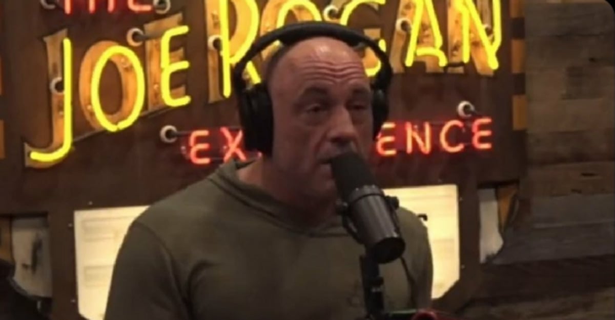 A man wearing headphones speaks into a microphone in a podcast studio, with a large illuminated "JOE ROGAN EXPERIENCE" sign visible in the background.