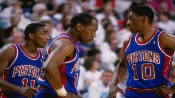 Three Detroit Pistons basketball players in blue jerseys, two facing each other in conversation while one stands nearby, with a blurred crowd in the background.