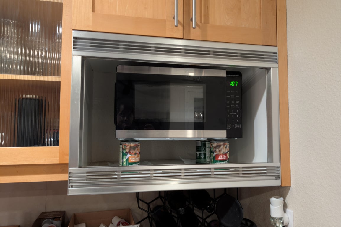 A microwave oven is installed in a kitchen cabinet, propped up by two canned food items underneath it. The microwave display shows the time 4:07. Wooden cabinets and shelves with glassware surround the area.