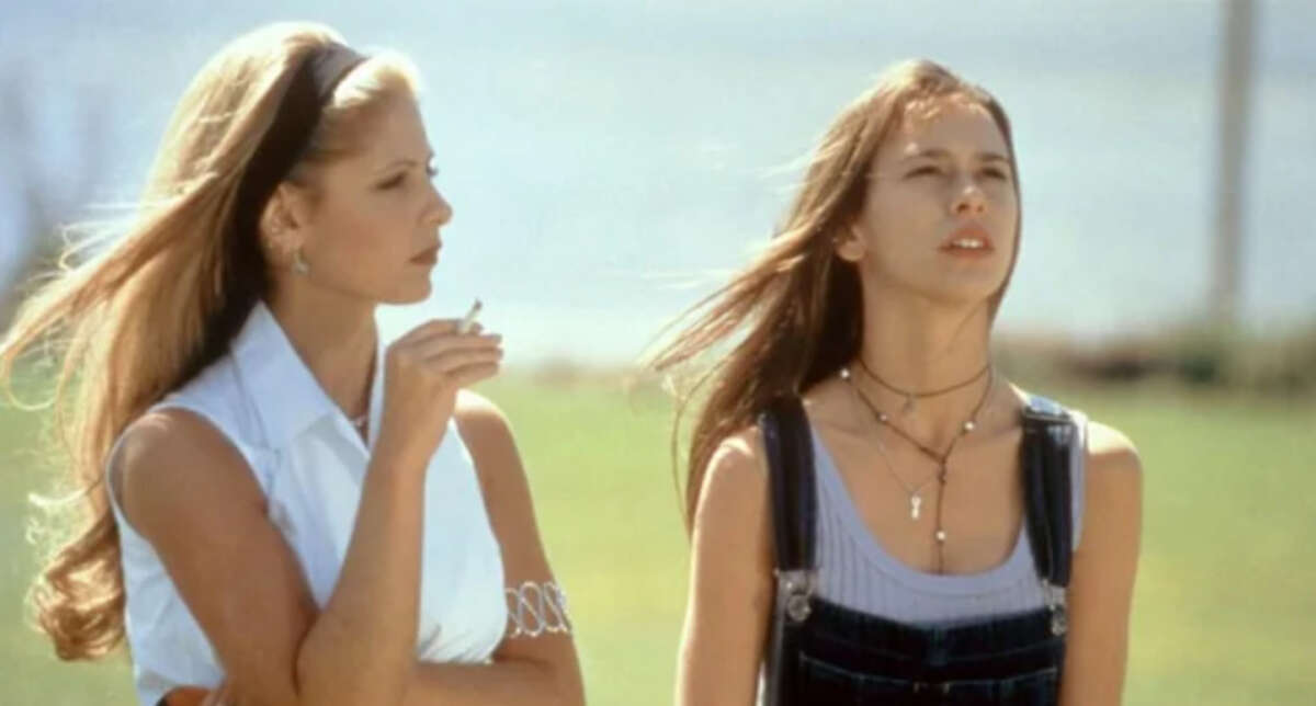 Two young women stand outdoors; one with long blonde hair holds a cigarette and looks at the other, who has long brown hair, wears overalls, and gazes upward. Both appear thoughtful under natural light.