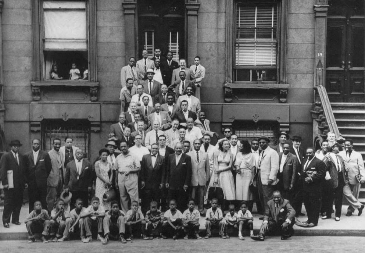 A large group of men, women, and children pose on the steps and sidewalk in front of apartment buildings, all dressed in suits, dresses, and hats, suggesting a formal gathering, with two people seen in the windows above.