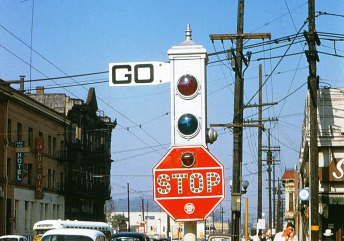 A vintage traffic signal with a large red stop sign and circular lights stands in the middle of a street. Above is a white “GO” sign. Old buildings, cars, and power lines are visible in the background under a blue sky.