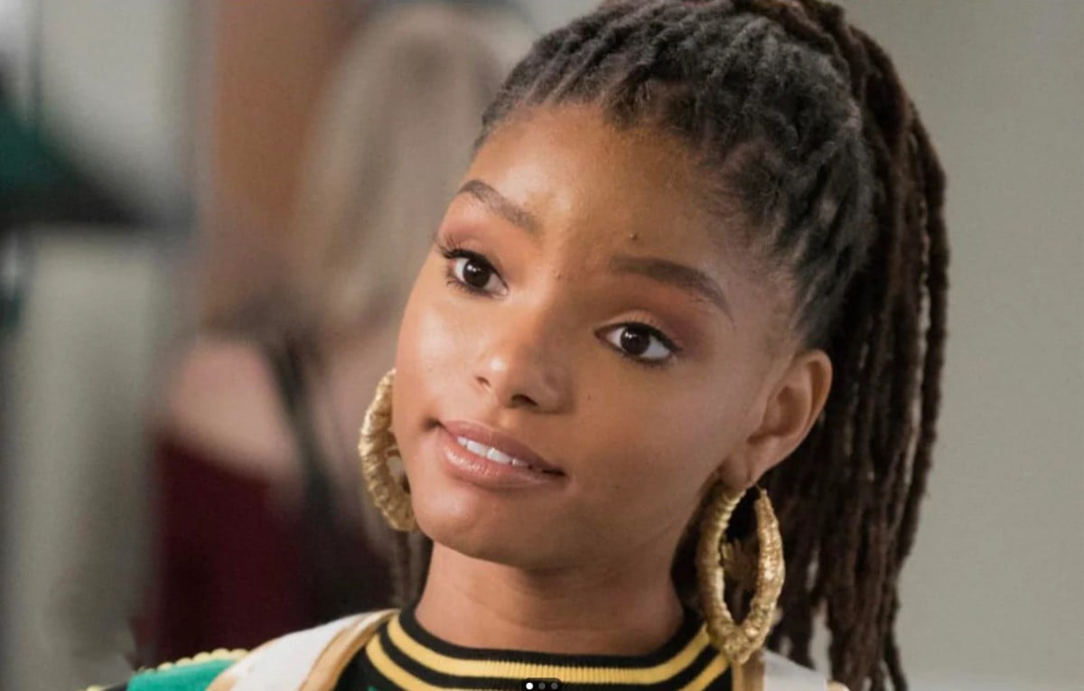 A young woman with long, styled dreadlocks and large gold hoop earrings looks slightly to the side, wearing a white top and a green-and-yellow jacket, with a calm and neutral expression.