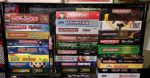 A shelf displays a large collection of Monopoly board games, including themed editions such as Pok&eacute;mon, Zelda, Fortnite, Fallout, Game of Thrones, Rick and Morty, and The Lord of the Rings.