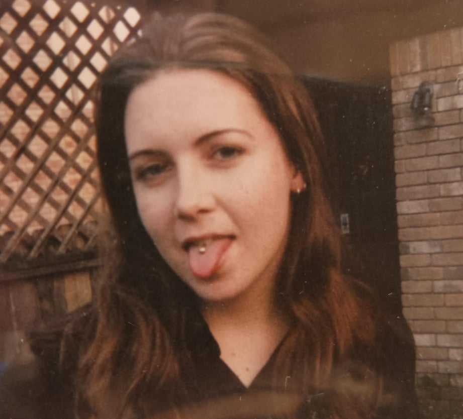 A young woman with long brown hair sticks out her tongue to show a tongue piercing. She stands in front of a wooden fence and lattice, with a brick wall and outdoor light fixture in the background.