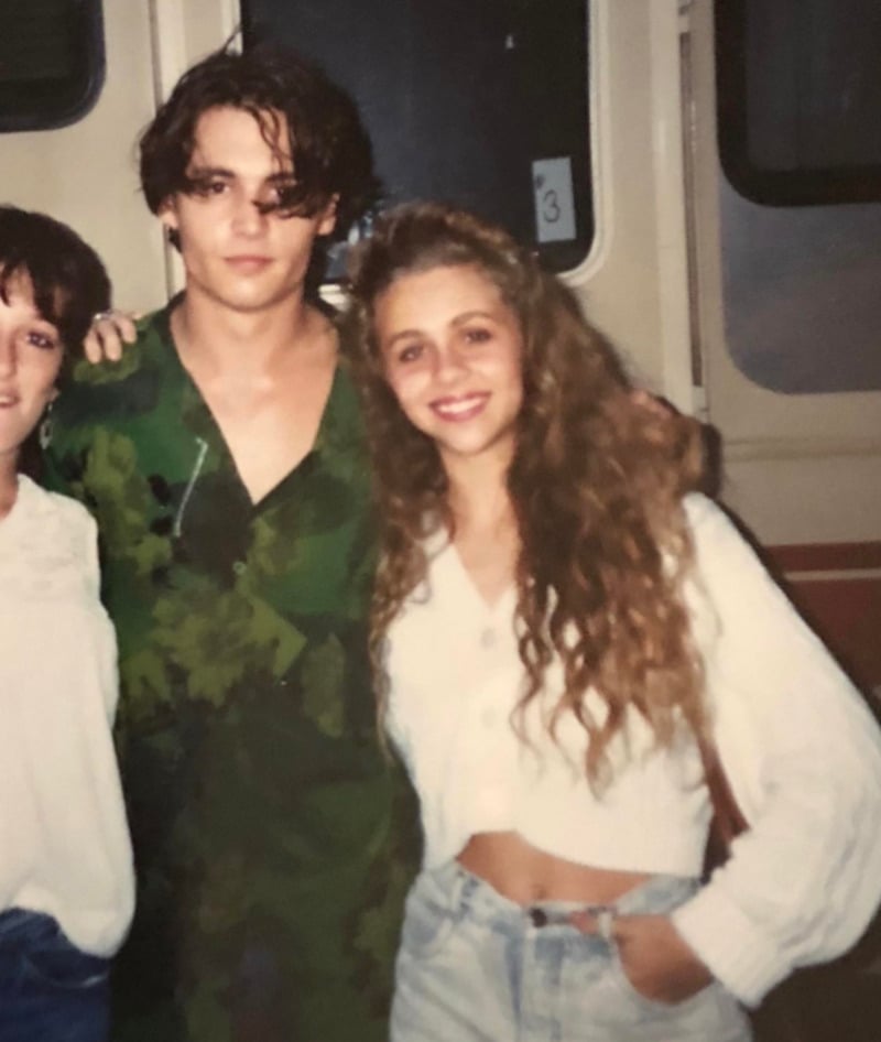 A young man in a green shirt stands next to a young woman with long, wavy hair wearing a white top and high-waisted jeans. Both are smiling, posing for a photo in front of a beige vehicle or trailer.