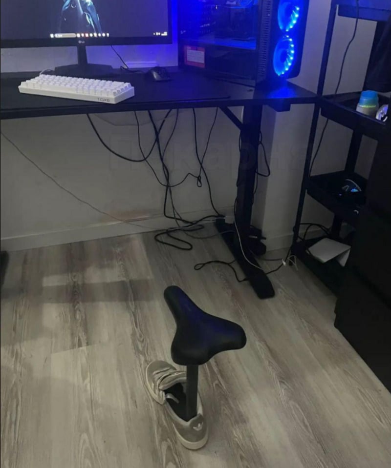 A minimalist desk setup with a computer and keyboard; below the desk is a bicycle seat attached to a sneaker, serving as an unconventional chair on a wood floor. Cables are visible under the desk.