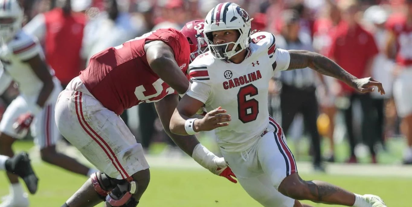 A South Carolina football player in a white uniform, number 6, runs with the ball as a defender in a red uniform attempts to tackle him during a daytime college football game.