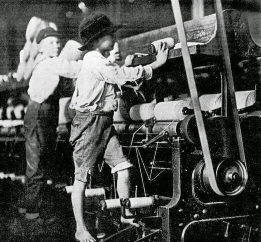 Two young boys, one barefoot, work at large textile machines in a factory. They wear old-fashioned clothes and hats, highlighting historical child labor conditions in an industrial setting.