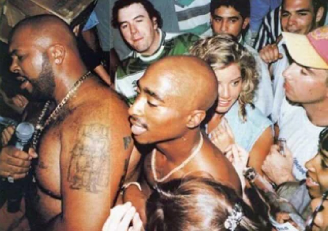 Two shirtless men, one holding a microphone, stand close together in a crowded room surrounded by people. The scene appears lively and energetic, with several people looking toward the camera.