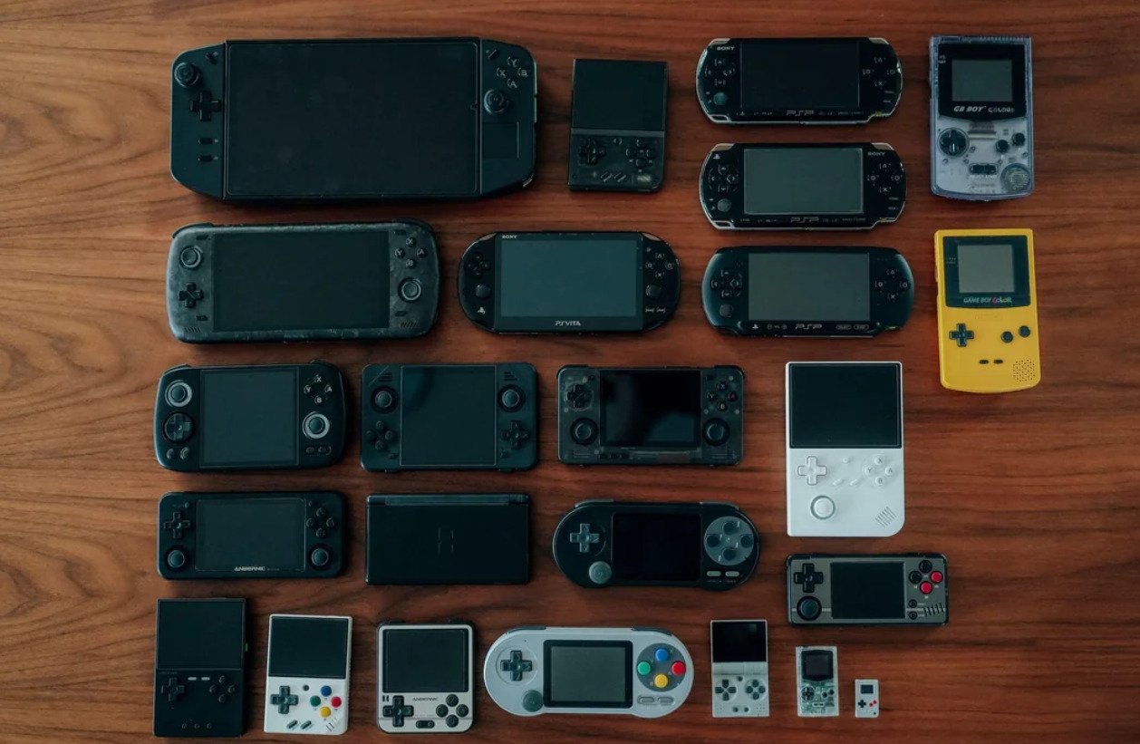 A collection of various handheld gaming consoles arranged neatly on a wooden surface, featuring devices of different sizes, colors, and styles, including both retro and modern models.