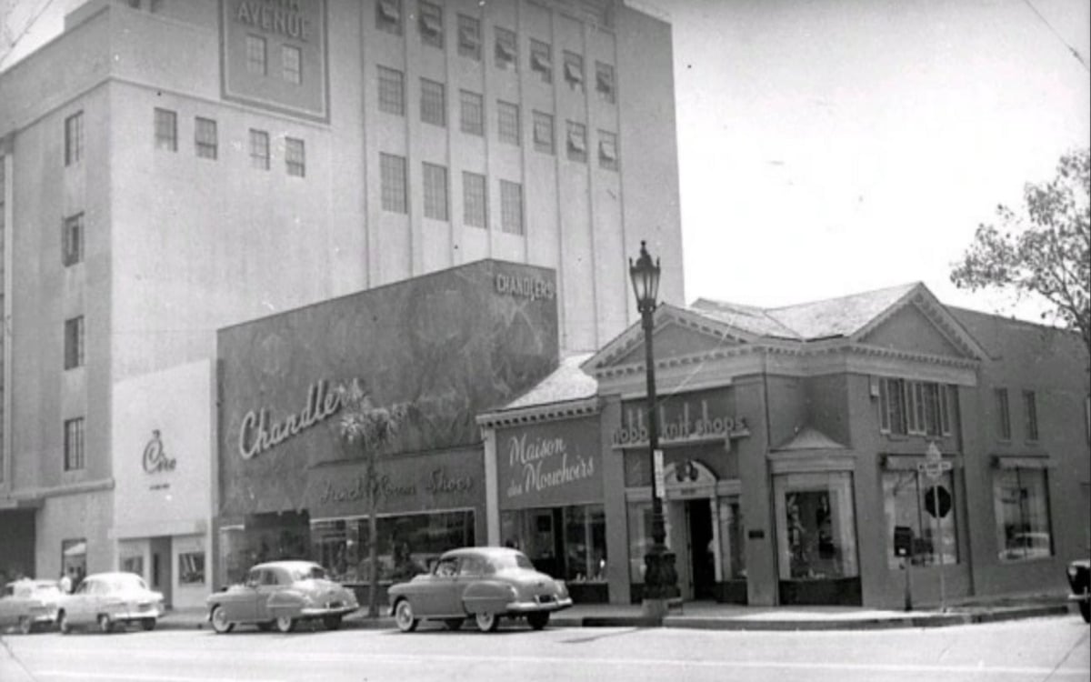 Black-and-white photo of a city street in the 1950s, showing vintage cars parked in front of storefronts, including Chandler’s Shoe Store and Maison de Manner’s, with a tall office building in the background.