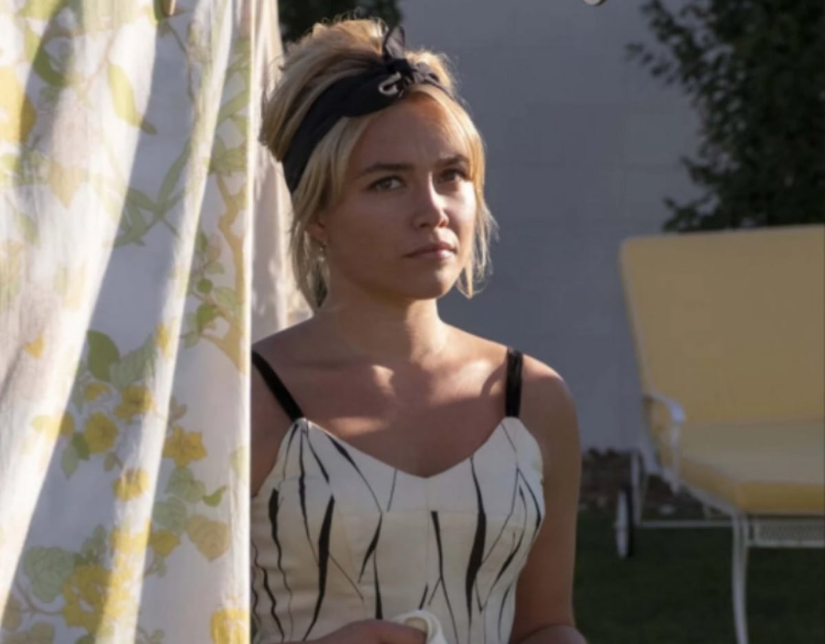 A woman with blonde hair in a black headband stands outdoors, holding a cup. She wears a sleeveless, patterned dress and looks thoughtful. A yellow floral curtain and a yellow lounge chair are in the background.