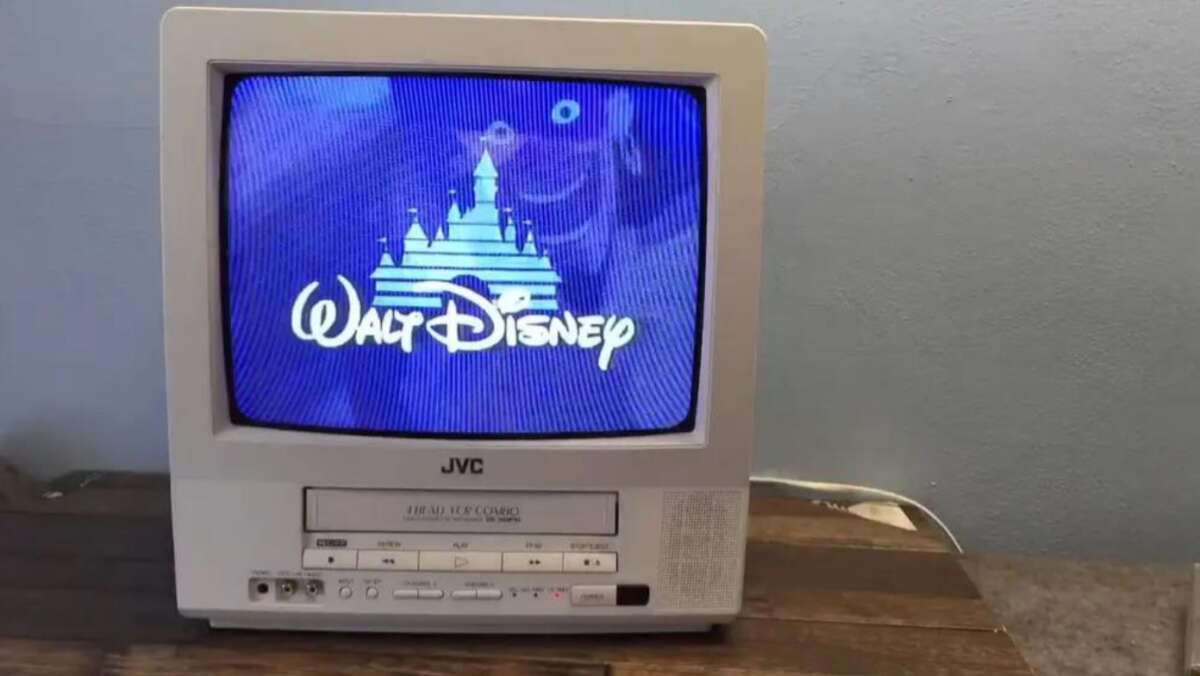 A JVC CRT television on a wooden surface displays the Walt Disney castle logo on its blue screen. The TV has visible buttons, a VHS slot, and sits against a plain light-blue wall.