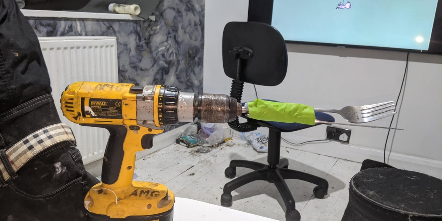 A yellow power drill has a fork taped to its chuck with green tape. The setup is on a white floor near a cluttered area with a black office chair, radiator, and a TV screen in the background.