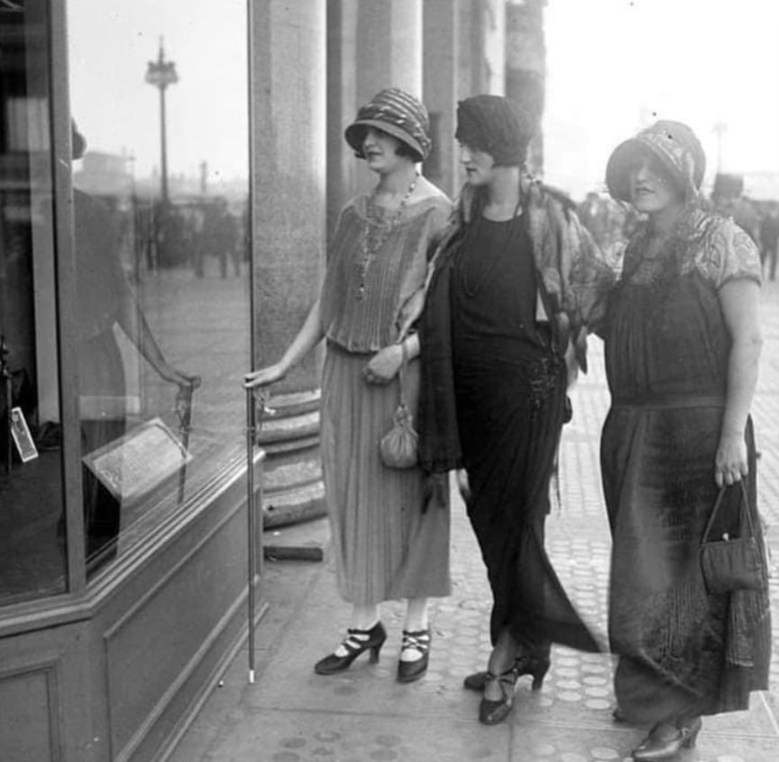 Three women in 1920s fashion, wearing cloche hats and dresses, stand on a city sidewalk looking into a shop window. The street and columns are visible in the background.