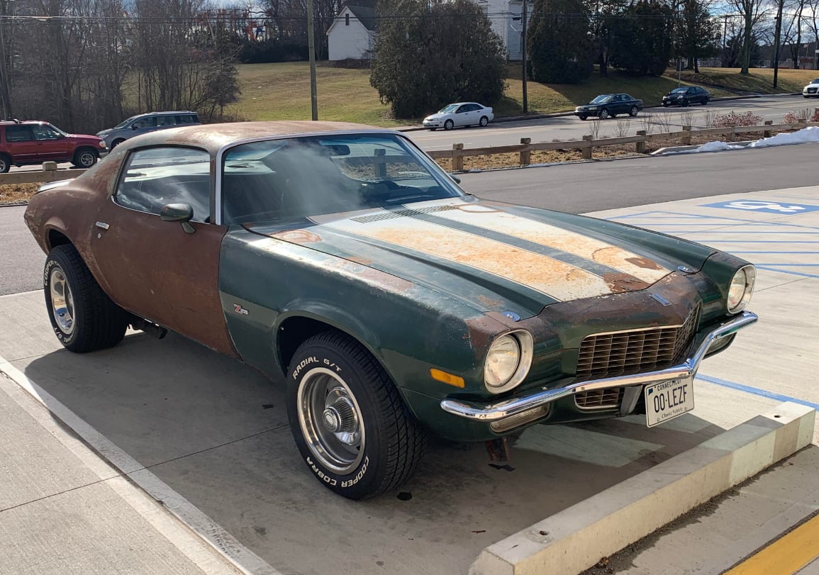 A green and brown vintage Chevrolet Camaro with rust and faded paint is parked in a lot on a sunny day. The car has white racing stripes and classic wheels.