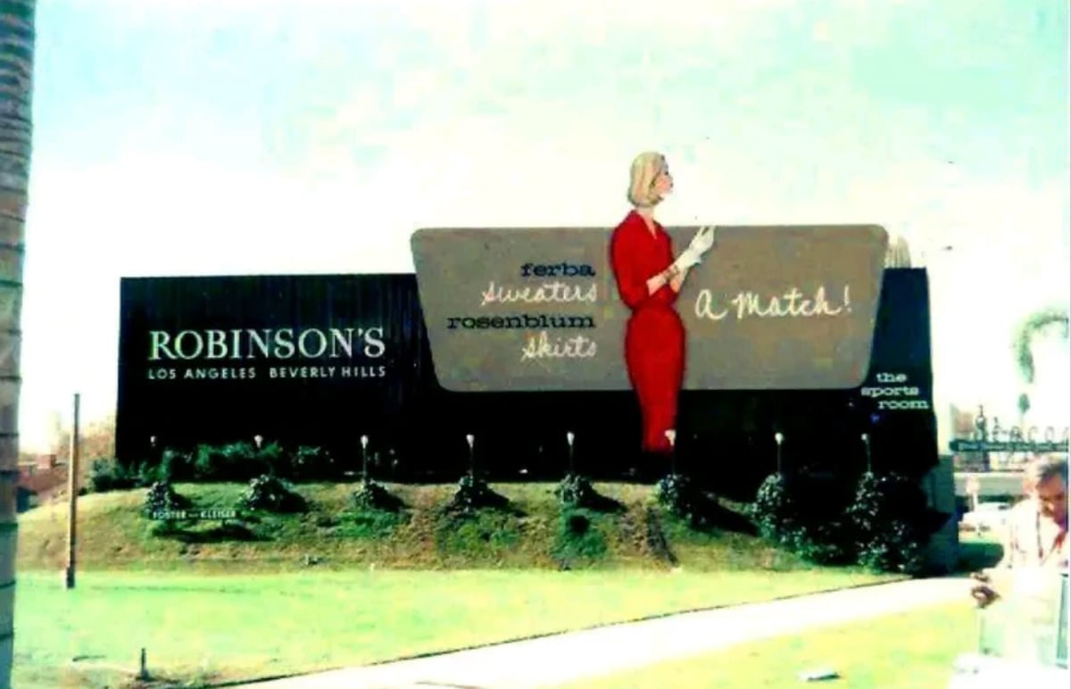 A vintage billboard for Robinson’s in Los Angeles and Beverly Hills features a woman in a red dress holding a card, with text advertising "Gerbe Sweaters" and "Rosenblum Skirts." Bushes and green grass are in the foreground.