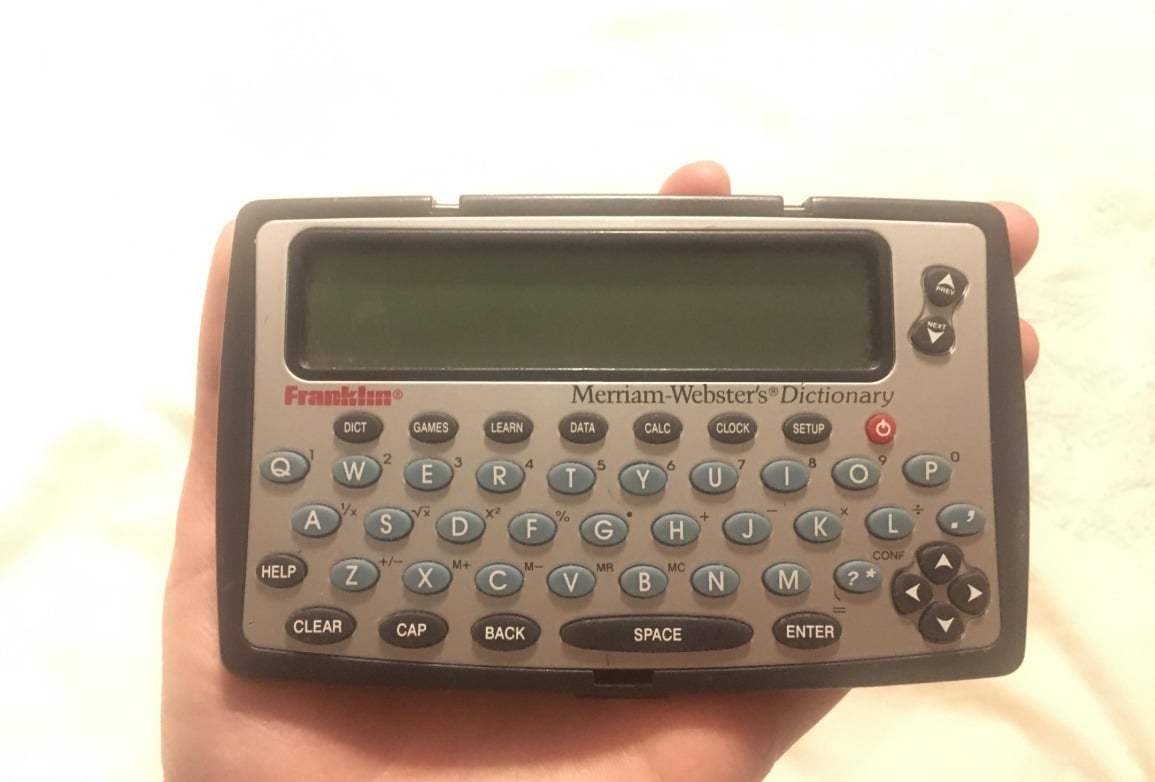 A hand holds a Franklin electronic device with a QWERTY keyboard and a large display screen. The device is labeled “Merriam-Webster’s Dictionary.”