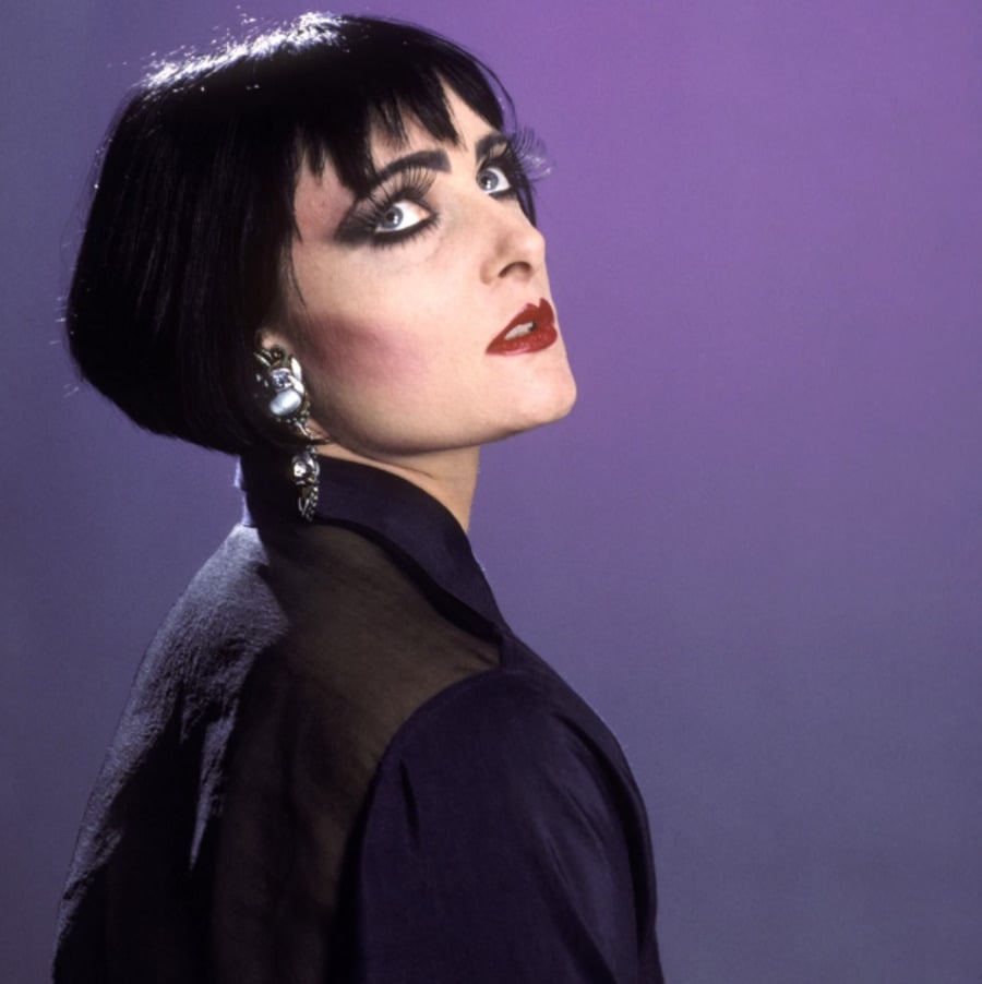 A person with short black hair, dramatic eye makeup, bold red lipstick, and silver earrings poses against a purple background, looking over their shoulder with a confident expression.