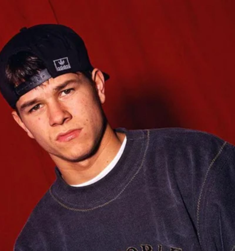 A young man wearing a dark sweatshirt and a black Adidas cap backwards stands in front of a plain red background, looking into the camera with a serious expression.