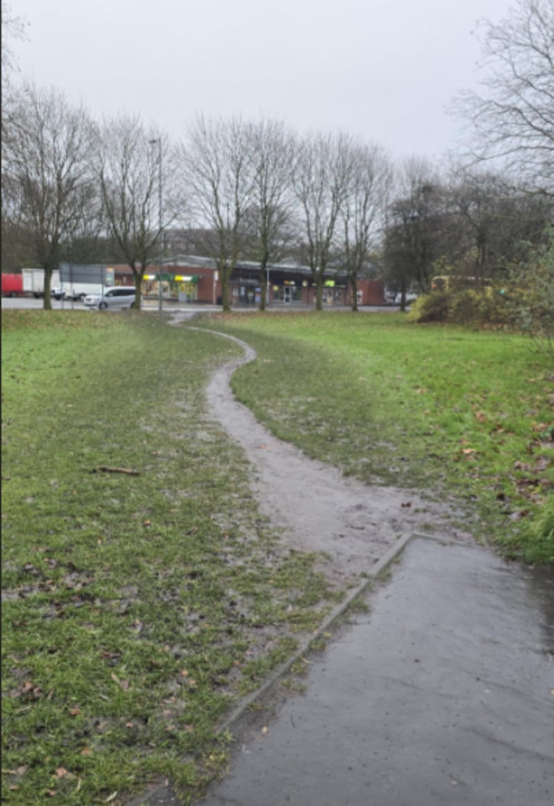 A muddy, narrow footpath cuts through a grassy field on a gray, rainy day, leading toward a row of shops and leafless trees in the background.
