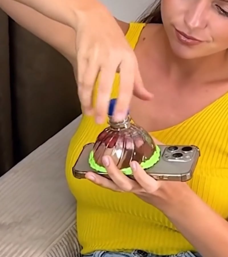 A woman in a yellow top holds a smartphone while using a suction device attached to the back of the phone, creating a seal with green material.