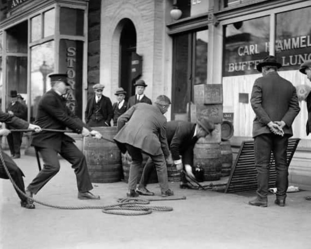 A group of men in suits pull a rope and lift barrels outside a storefront on a city sidewalk, while several others stand and watch. The scene appears to be from the early 20th century.