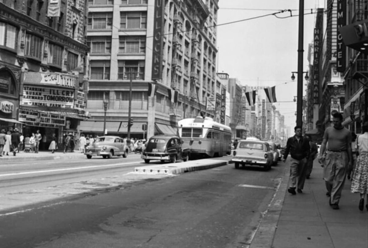 Black and white photo of a busy city street in the mid-20th century with cars, a trolley, pedestrians, and tall buildings with storefronts and marquee signs.