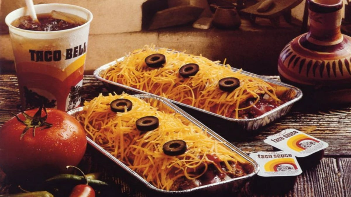 A vintage Taco Bell meal with two trays of enchiladas topped with shredded cheese and sliced olives, a soft drink, taco sauce packets, a tomato, and chili peppers on a rustic table.